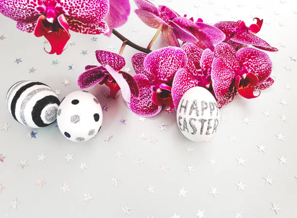 Composition of glitter inscription Happy Easter on a white egg and metallic stars spangle sequins. Red orchid, eggs with black and silver lines and polka dots. Flat lay, close up, copy space
