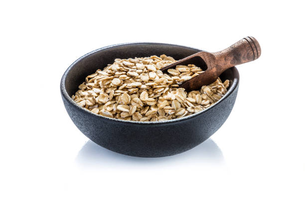Front view of a black bowl full of uncooked oat flakes and a wooden serving scoop isolated on white background.