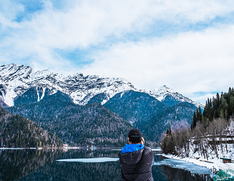 A man in a black jacket with a blue hood takes a picture of a beautiful winter landscape of the Caucasus mountains and Lake Ritsa, located in Abkhazia