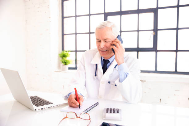 médecin masculin aîné utilisant le téléphone mobile et l’ordinateur portatif tout en travaillant dans le bureau du docteur - telephone keypad old white photos et images de collection