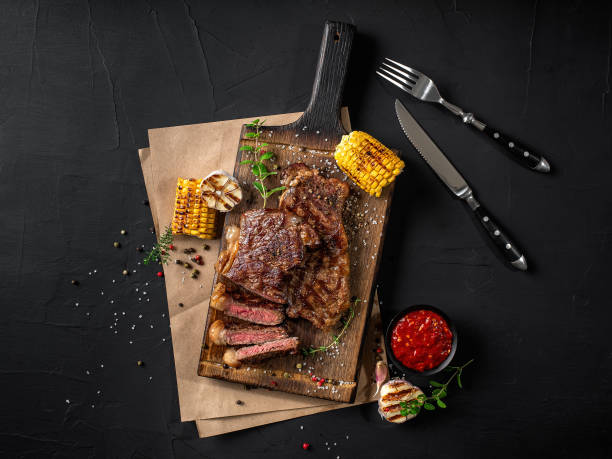 Fried steaks with herbs and spices on wooden board, parchment, grilled garlic, corn, sauce, fork and knife on black background. Close-up. Top view
