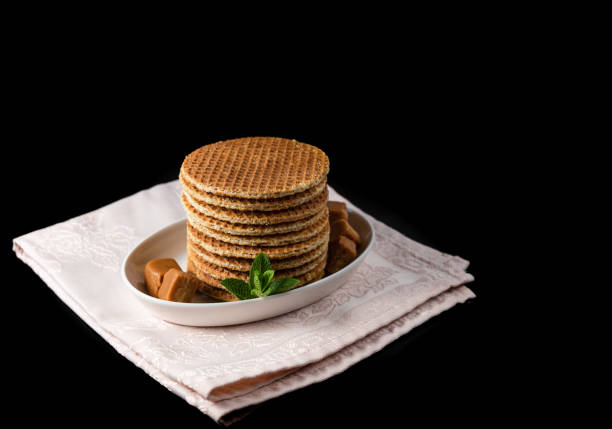 Several stacked stroopwafels or dutch waffles made from caramel syrup, delicate decoration with some coffe candies and mint leaves, on a black background