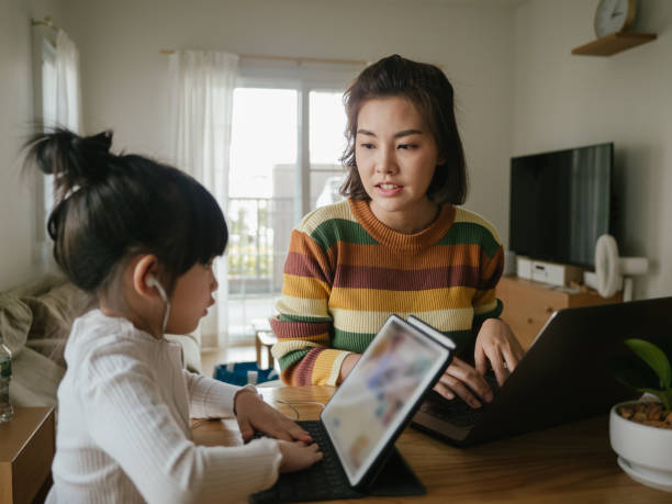 Young mother help little girl with homework at home. Asian girl doing homework on tablet while young woman is using her laptop at home. asian parent cellphone stock pictures, royalty-free photos & images