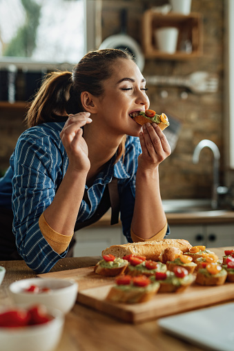 Young woman enjoying in taste on healthy bruschetta with her eyes closed in the kitchen.