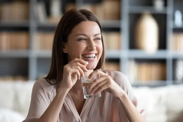 Photo of Smiling young woman holding pill and glass of pure water.