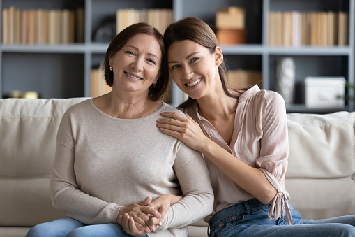 Portrait of happy grownup daughter touching shoulder of pleasant smiling old mature mother, relaxing together on couch. Affectionate two generations family looking at camera, posing for photo at home.