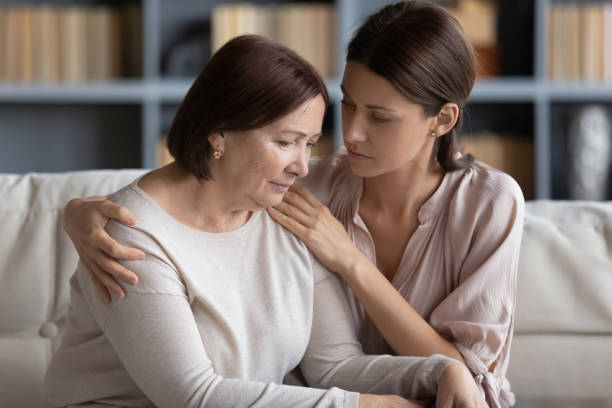 Compassionate woman embracing shoulder of unhappy stressed middle aged mommy. Young compassionate woman embracing shoulder of unhappy stressed middle aged mommy, comforting her at home. Kind grownup daughter supporting caring of older mature mother, sitting together on couch. mercy hug stock pictures, royalty-free photos & images
