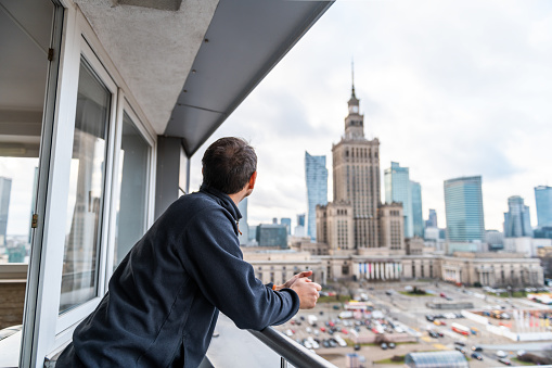 Young man standing looking in apartment with view of Warsaw, Poland famous Palace of Science and Culture cityscape building on balcony