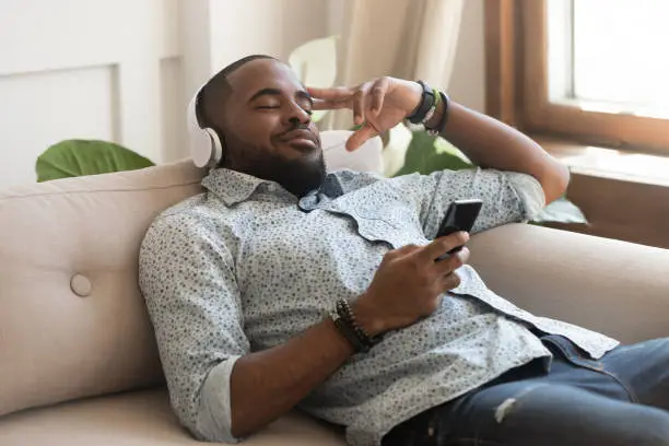 Photo of African guy holding smartphone closed eyes listen music on headphones