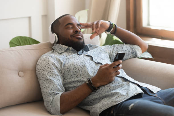 african guy holding smartphone closed eyes listen music on headphones - cair no sofá imagens e fotografias de stock