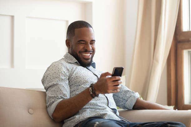 istock Cheerful african guy sitting on couch spending time using smartphone 1217477770