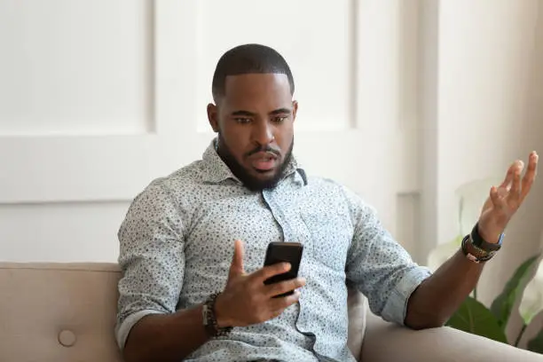 African astonished man sitting on sofa holds cellphone read e-mail sms feels shocked received terrified news, guy looks at online calendar forgot missed important meeting, phone crash problems concept