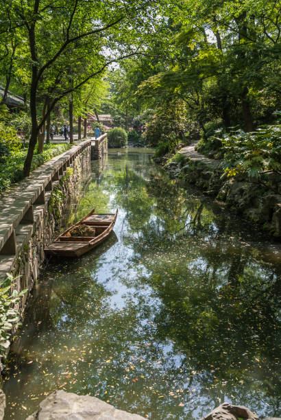 Small sloop on canal at Humble Administrators garden, Suzhou, China. Suzhou China - May 3, 2010: Humble Administrators Garden. Small wooden sloop sits on reflecting greenish canal with green foliage on both sides. People in distance. suzhou creek stock pictures, royalty-free photos & images