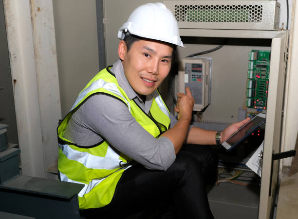 l’ingénieur électricien pointe vers le générateur d’alimentation et la tablette de fixation à la salle de commande de l’usine. port d’un gilet de sécurité et d’un casque blanc - control panel power switch video game photos et images de collection