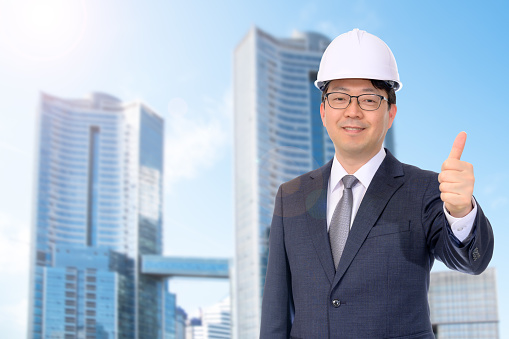 A middle-aged Asian businessman smiles in a white hard hat in front of a modern building.