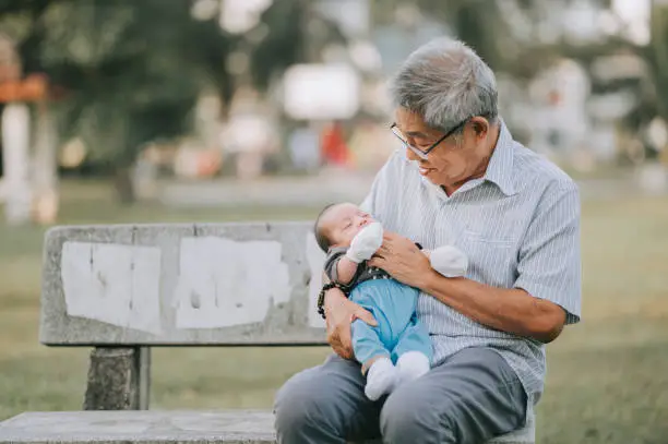 Photo of an asian chinese senior man grandfather looking at his grandson in the public park sitting on the bench while the baby sleeping