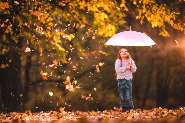Happy little girl holding an umbrella while autumn leaves are falling onto her at the park. Copy space.