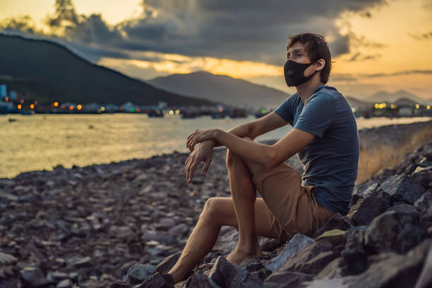 Self-isolation in nature. Young man in Fashionable black medical mask with filter sitting by the river. Coronavirus 2019-ncov epidemic concept. Man in a black medical mask. Portrait of a man with expressive eyes during a virus or disease epidemic of Coronovirus. autoaislamiento en la naturaleza. joven en máscara médica negra de moda con filtro sentado junto al río. concepto epidémico coronavirus 2019-ncov. retrato de un hombre con ojos expresivos durante una epidemia de virus o enfermedades de coron