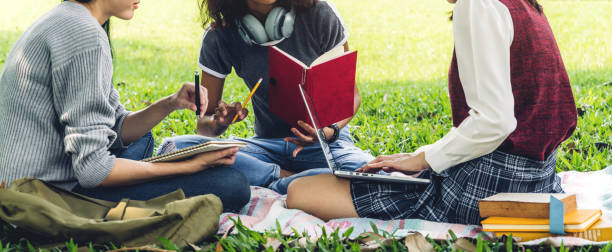 istock Group of smiling international students or teenagers sitting and using laptop computer doing their homework with book together in park at university.Education and friendship Concept 1217528610