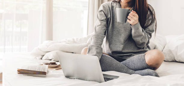 Young woman relaxing and drinking cup of hot coffee or tea using laptop computer on a cold winter day in the bedroom.woman checking social apps and working.Communication and technology concept stock photo
