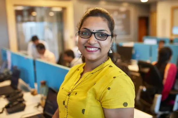 Three-quarter front view of confident mid adult Indian female office worker wearing glasses, yellow traditional blouse, and smiling at camera.