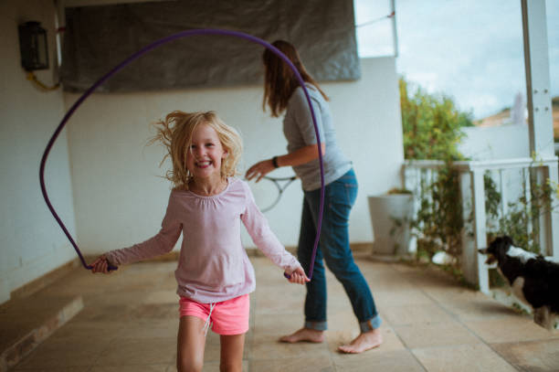 Front view Skipping rope at home with mom during lockdown Mother and daughter time together at home doing exercises having fun keeping busy during lockdown.  The cute 6-7-year-old blond girl is skipping rope and jumping having fun on the front stoop.  A pet Border Collie dog is watching the ball of the woman playing tennis against the wall. jump rope stock pictures, royalty-free photos & images