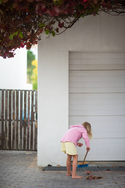 Child Picking up leaves home chores during lockdown outdoors.  She is using a broom to rake the leaves onto a heap in front of a garage door.