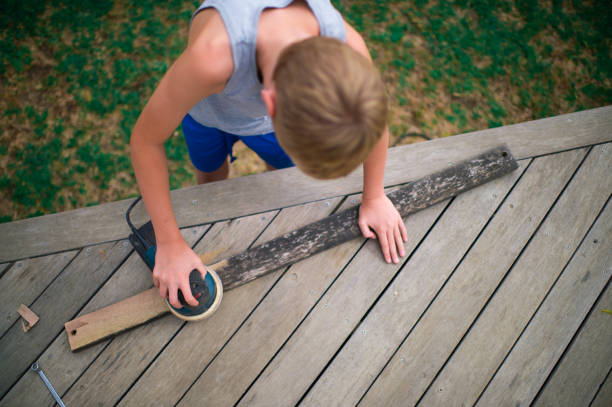 A young blond hair Boy is carefully using an orbit sander to clean a wooden plank from a bench he is renovating.  He is sanding the wooden piece of wood down.  It is an electrical orbit sander with a blue colour. He is using a wooden deck as a workbench in the backyard.