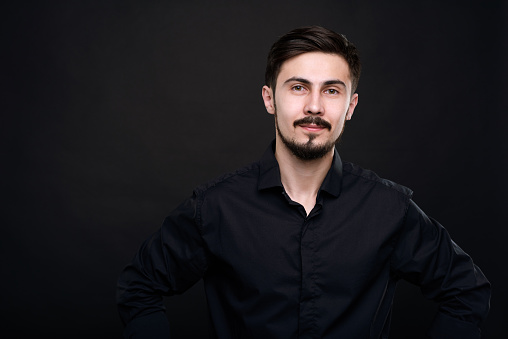 Portrait of content handsome young man with mustache and beard standing against black background