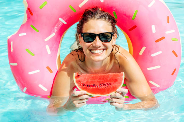 счастливая женщина в солнцезащитных очках с красным арбузом наслаждается в бассейне с розовым резиновым кольцом - swimming pool women floating on water bikini стоковые фото и изображения