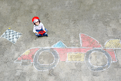 Happy healthy little boy having fun with race care picture drawing with chalk. Creative leisure for children. Adorable kid child sitting on asphalt and creating with colorful chalks