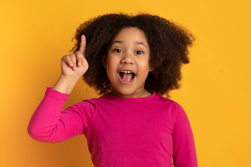 I Have Idea. Little african american girl pointing finger up and looking at camera, standing over yellow studio background with free space