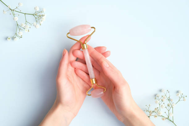 mano femenina sosteniendo rodillo de masaje facial de cuarzo rosa sobre fondo azul con flores de gypsophila. masajeador facial con piedra de jade, antienvejecimiento, herramienta antiarrugas de cuidado de la piel de belleza - health spa women spa treatment massager fotografías e imágenes de stock