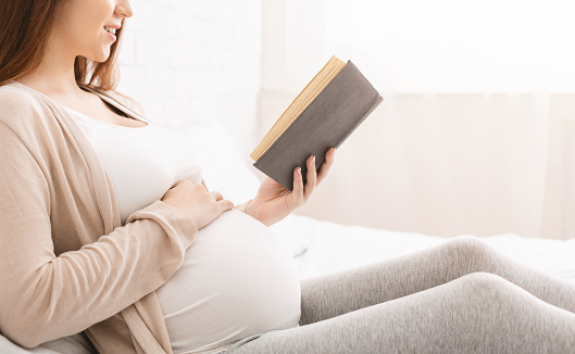 Stay at home concept. Smiling pregnant woman reading her favorite book, sitting on bed, free space