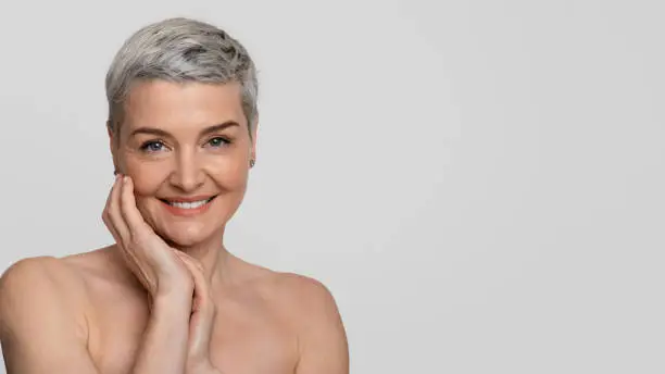 Mature Beauty. Portrait of charming nude middle-aged woman touching her skin and smiling at camera, light background, panorama with empty space