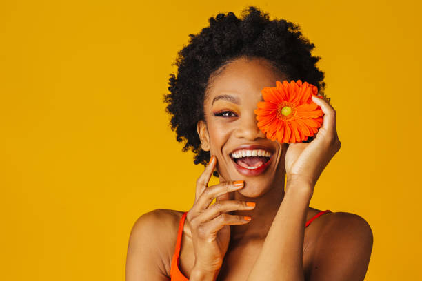 портрет счастливой возбужденной молодой женщины, держащей оранжевую маргаритку герберы, закрываюющую ее глаз - human face smiling spring women стоковые фото и изображения