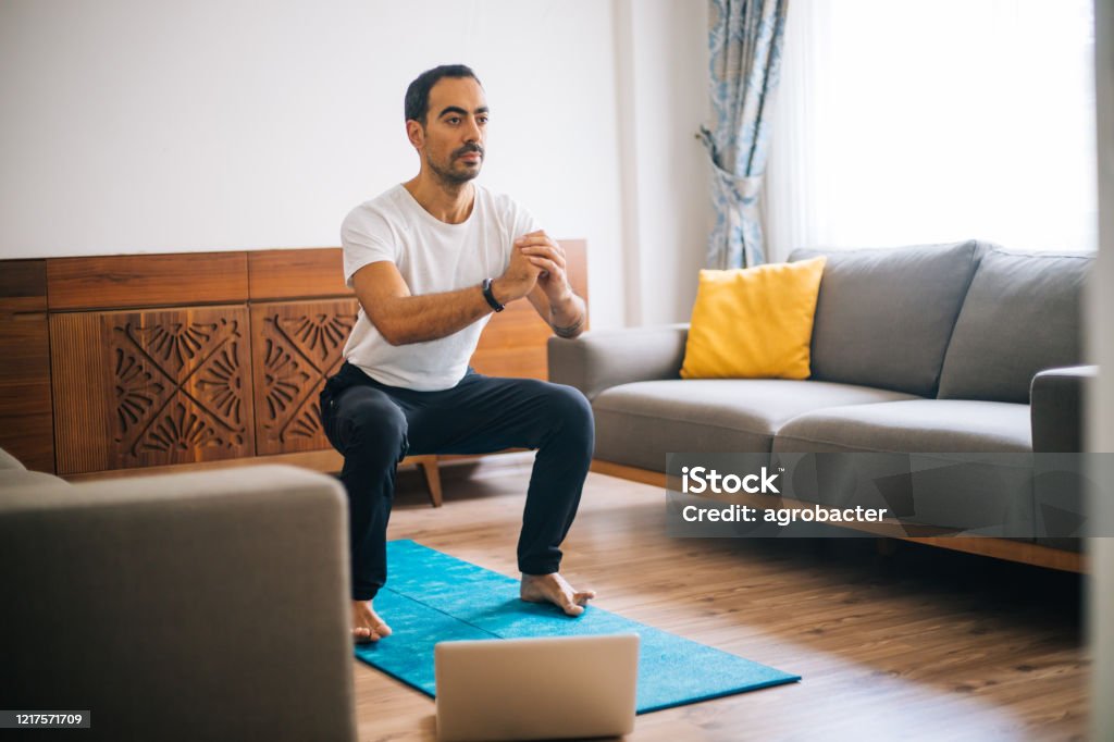 Muscular Athletic Fit Man in T-shirt and Shorts is Doing Squat Exercises at Home Squatting Position Stock Photo