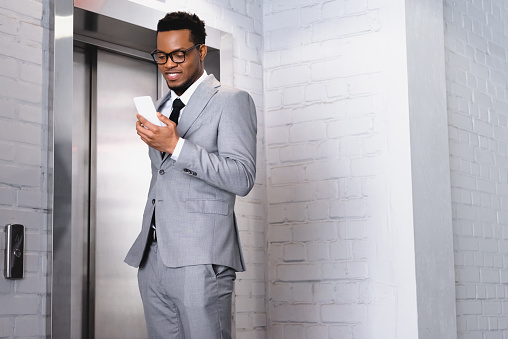 smiling african american businessman holding smartphone while standing near elevator