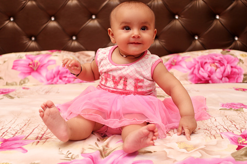 Beautiful innocent 6 months baby in pink frock sitting on bed and playing lonely in living room.