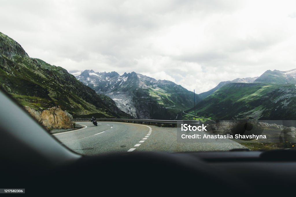View from the car of the  dramatic mountain pass by car in Switzerland Have a great road trip at the mountains of Switzerland - driving at the scenic mountain road with view of the peaks and glacier during cloudy summer day at Furka Pass Car Stock Photo