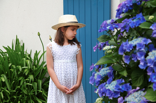 Young girl in white dress stay near old cottage with blue colour door. Bright Hydrangea with blurred blue flowers.