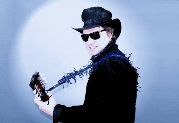 Cool country singer and guitarist in a hat and in sunglasses playing the acoustic guitar on a blue backdrop. stock photo