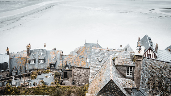 View of the bay of Mont Saint-Michel from a terrace in the old town\nThe weather is very cloudy\nIn the foreground we see a garden located on a terrace in the middle of the old town and its dwellings\nOn the left is the city cemetery