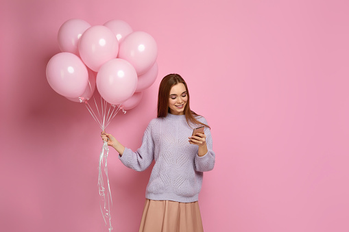 beautiful smiling caucasian woman in knitted sweater with pastel pink air balloons using smart phone on pink background. copy space