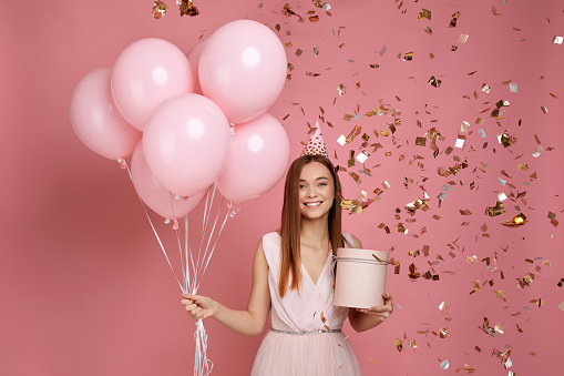Beautiful smiling woman in dress and birthday hat holding gift box and pastel pink air balloons and falling confetti against pink background. cute happy young girl celebrating birthday party.