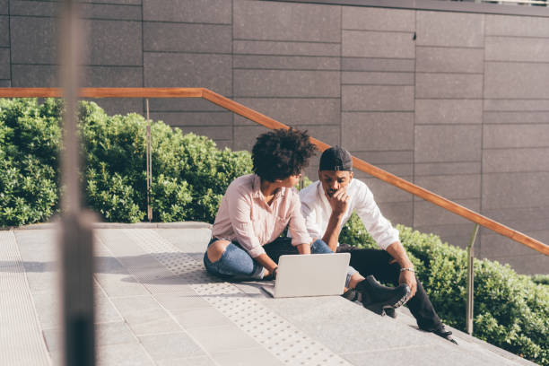istock Young multiethnic couple using computer sitting outdoor in the city 1217605286
