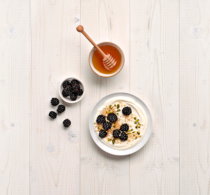 Bowl with yogurt, blackberries and honey, viewed from above on a light wood background.