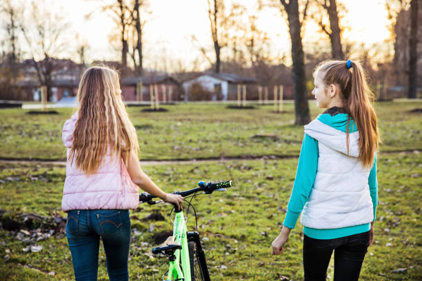 Fraternal twin sisters walking at the park together, one is leading a bicycle