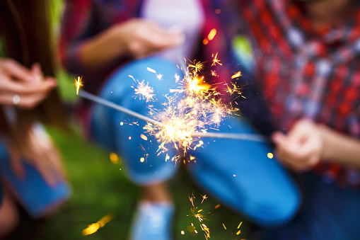 Group of friends having fun with sparklers sitting on grass in summer park. Teenagers having fun together. Group of young people enjoying party. Rest, fun, summer, holidays concept.