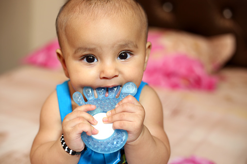 Close-Up portrait of cute Indian baby girl sitting on bed and she chewing on sterilized water filled teether.